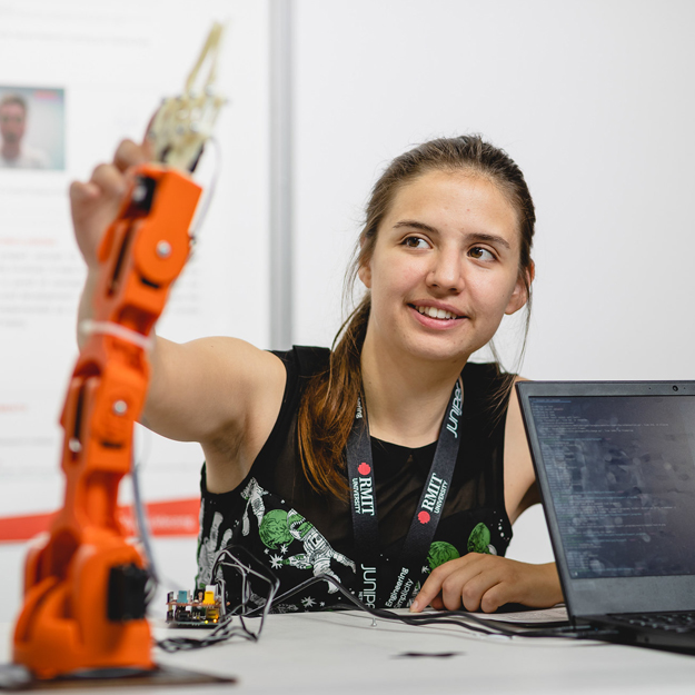 A girl with her hair in a ponytail wearing an RMIT lanyard, sitting with a laptop in front of her, reaching out with one arm to adjust something on an orange mechanical arm.