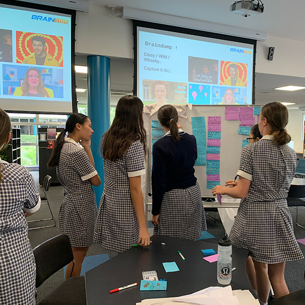 Five school girls watch a presentation on a projector while organising sticky notes on a whiteboard.