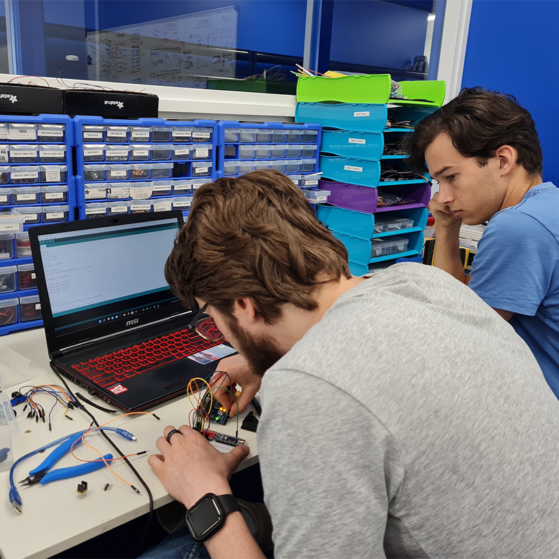 Two men in an engineering lab sit next to each other. The one on the left in a grey t-shirt works on a circuit board, and the one on the right supervises.