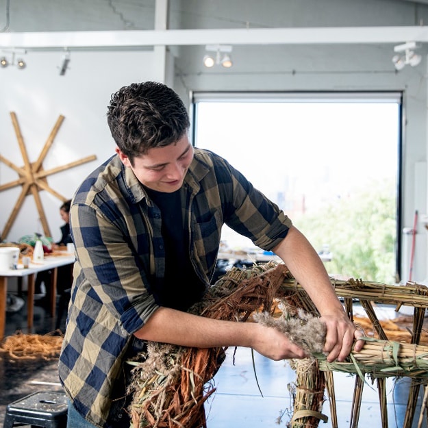 A man with dark hair, wearing a blue and olive tartan shirt, works away constructing something out of wood and fibres in a workshop.