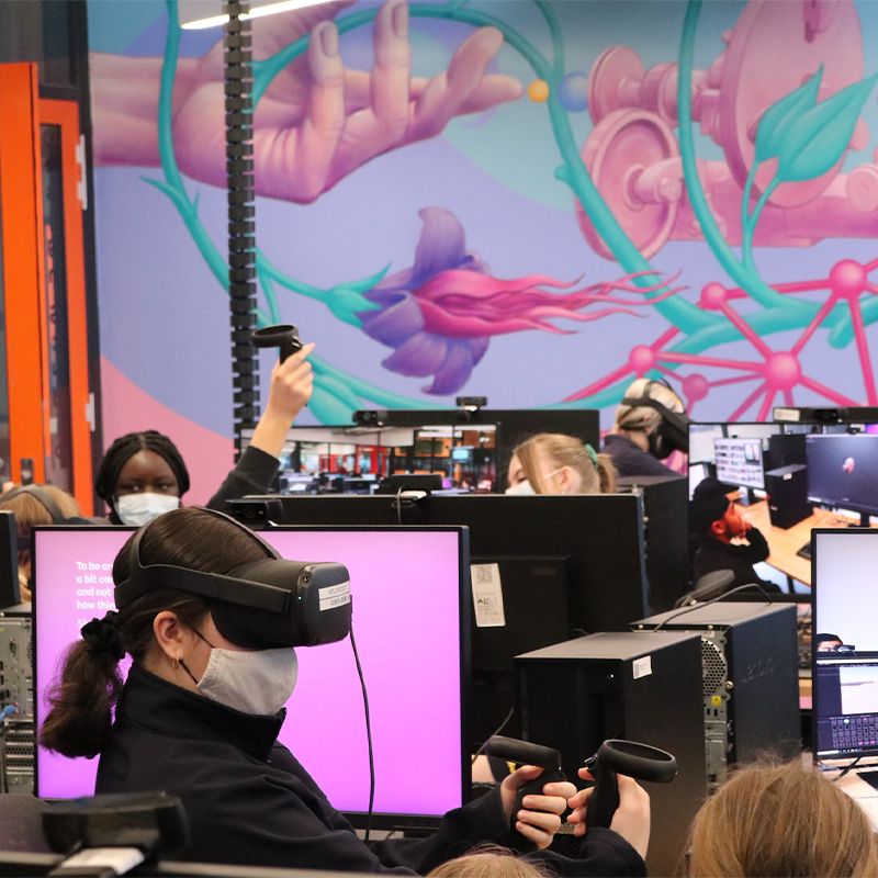 A computer lab at Wyndham Tech School, with flowers painted on the walls, filled with many computers, each person at a computer wearing a VR headset and testing state-of-the-art software.