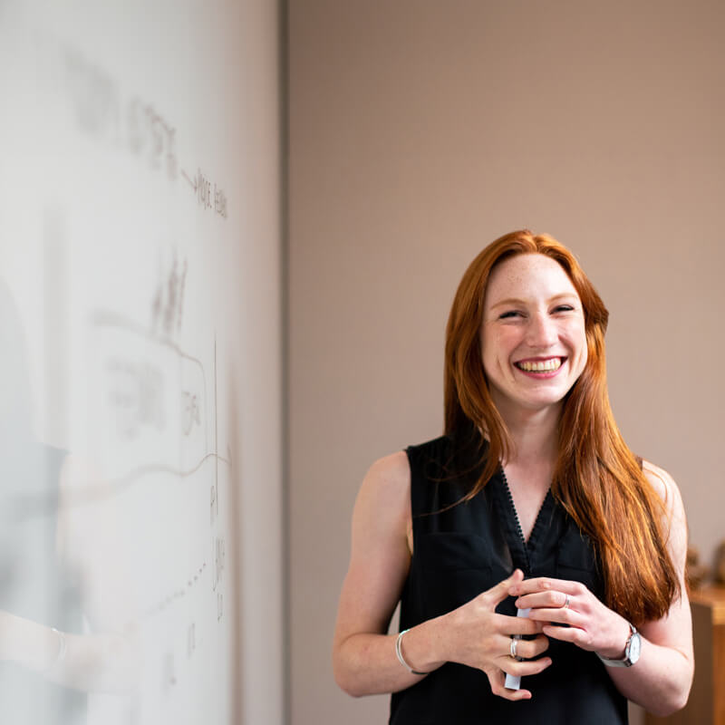 A youthful woman with long, healthy orange hair and freckles, smiling happily, stands next to a whiteboard wall with annotations scribbled across its surface.
