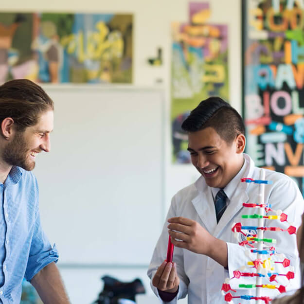 A teacher man with his hair tied back into a bun, working with a scientist in a white lab coat, laughing together in a school science lab environment.