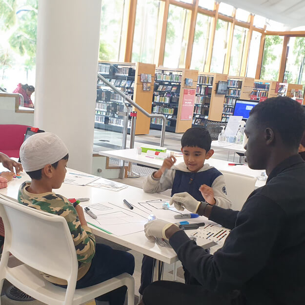 A man in white scientific gloves educates two young boys about science at a table in a brightly lit school library.