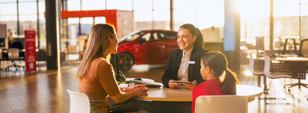 A woman in a blazer with a name badge sits at a round table smiling at a woman and a young girl inside a Toyota dealership.