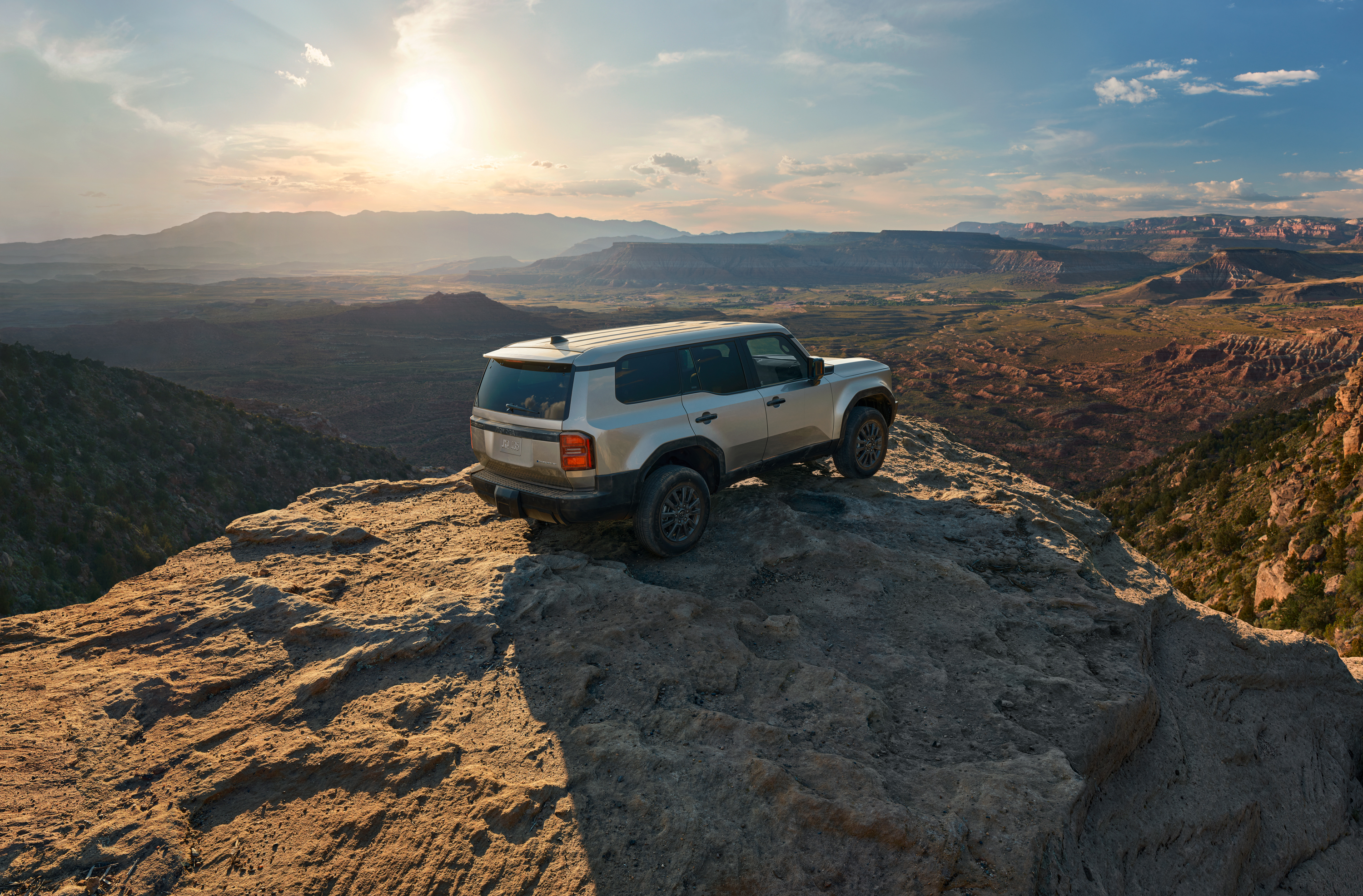 A silver Toyota LandCruiser Prado is parked on top of a rocky peak overlooking an Australian a red rocky landscape of cliffs and canyons. The sun has lowered and is beginning to set, a scattering of wispy clouds appear in the blue sky.