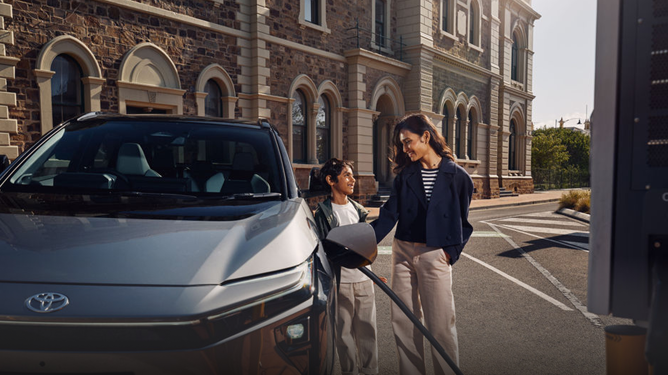 A woman and a child stand beside a gray electric bZ4X that’s plugged in and charging on a city street. They smile at each other while the car charges, with a stone heritage building in the background.