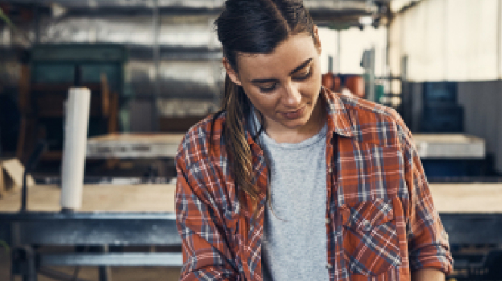 A woman wearing a red flannel shirt and a grey tee, hair tied back, examining paperwork and a calculator in a large tin shed.