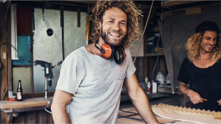 A man smiling, leaning against a wooden workbench, with noise-cancelling construction grade earmuffs hanging around his neck