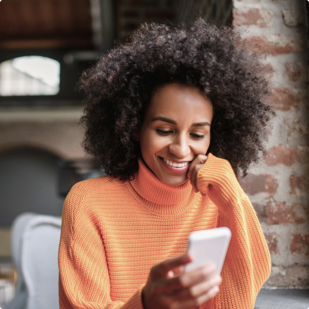 A woman sits indoors, smiling as she looks down at the screen of the mobile phone she’s holding.