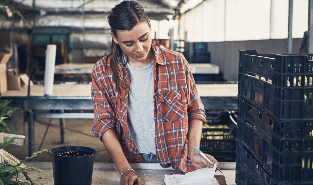 A woman wearing a red flannel shirt and a grey tee, hair tied back, examining paperwork and a calculator in a large tin shed.