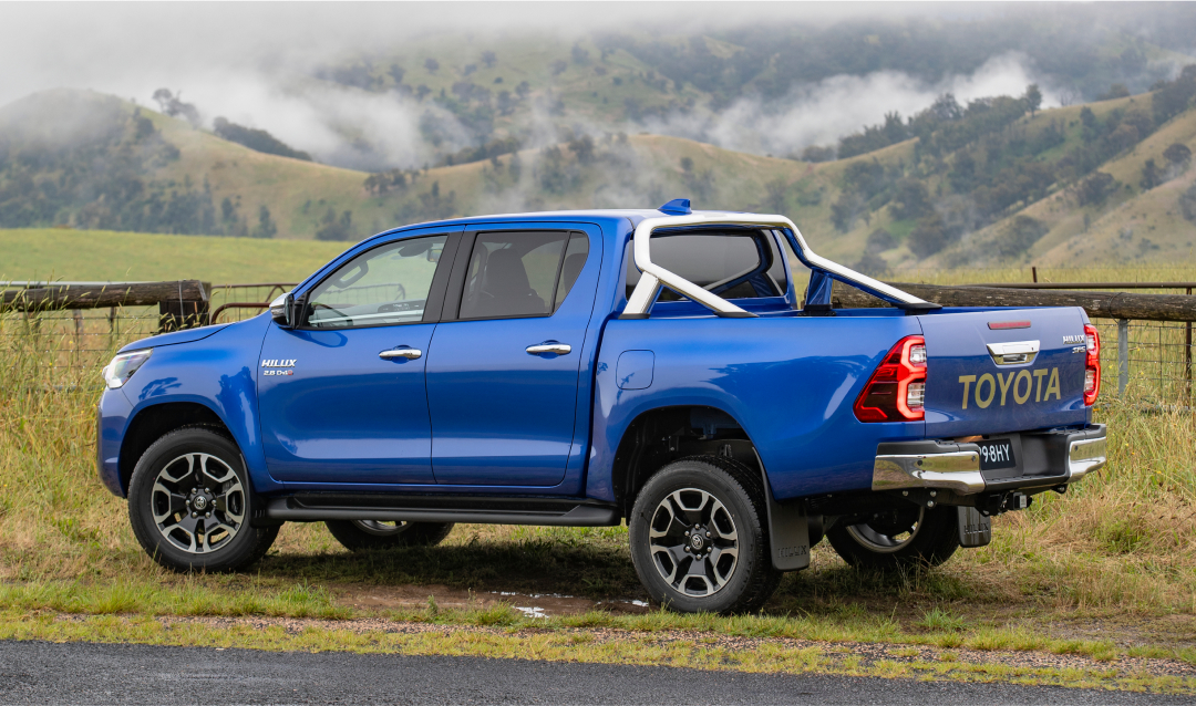 A blue Toyota HiLux parked in front of a wooden fence, overlooking a green misty valley.
