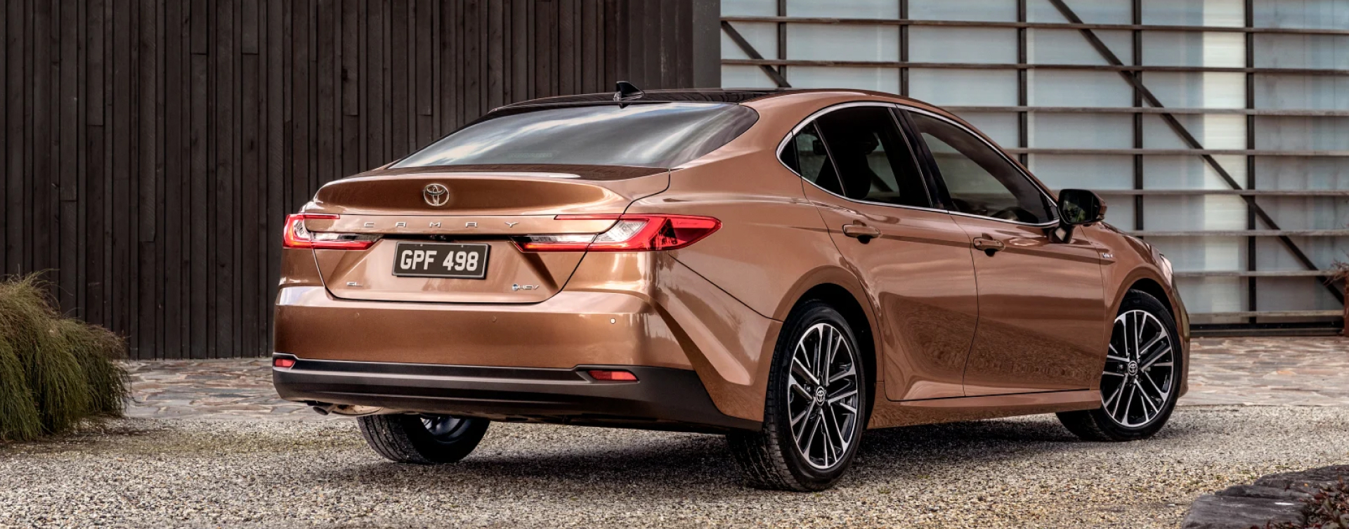 A bronze Toyota Camry Sedan parked on a modern driveway in front of a wooden building.