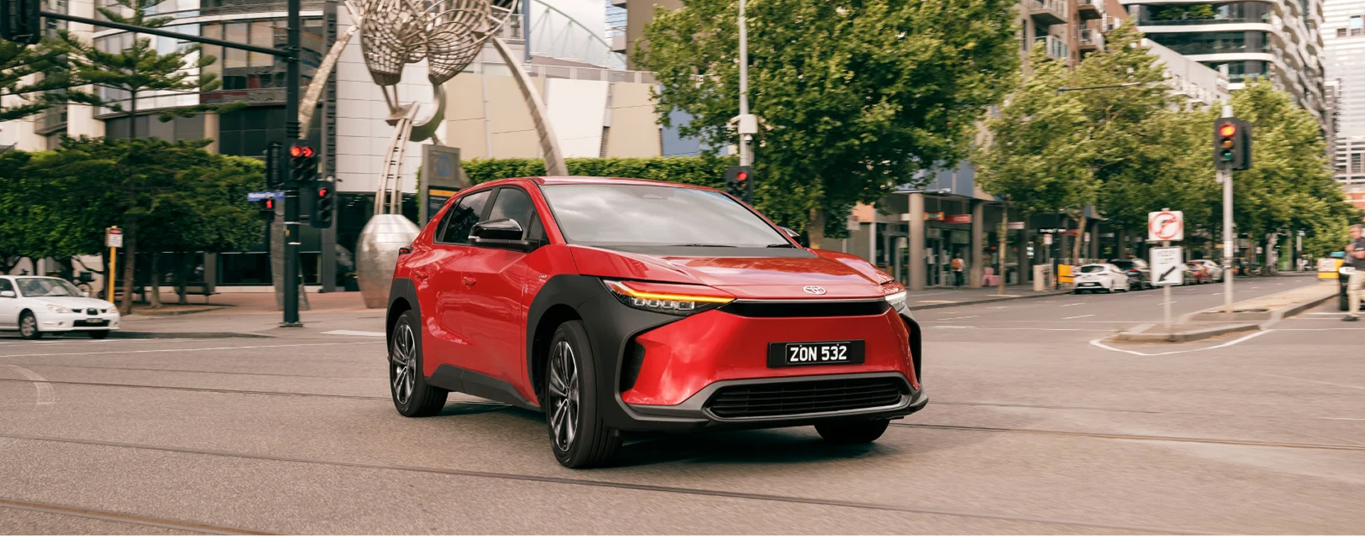 A red Toyota bZ4X crosses a quiet intersection in Melbourne’s CBD, the street lined with trees, and an artistic sculpture in the background. 