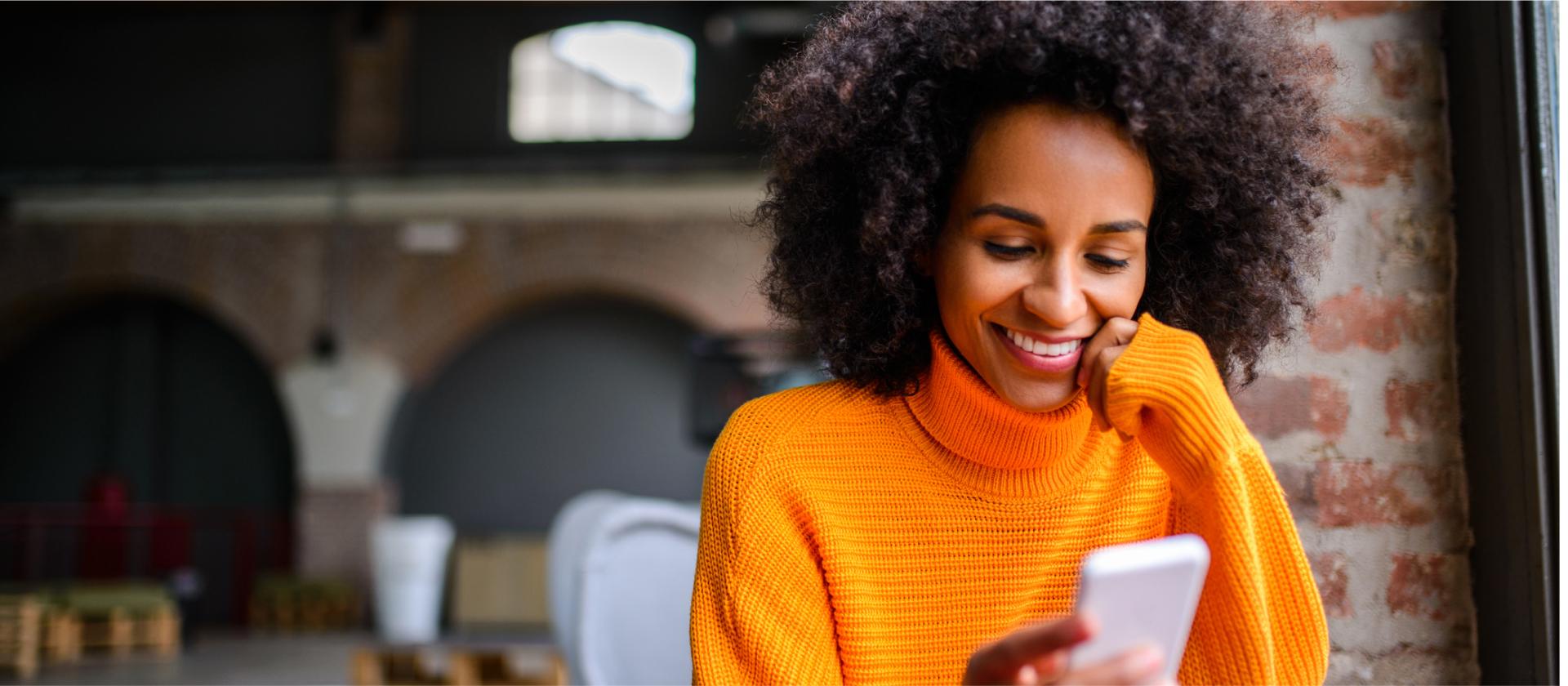 A woman in a bright orange jumper, sitting in a cafe with exposed brick walls, smiling as she looks down at her smartphone.