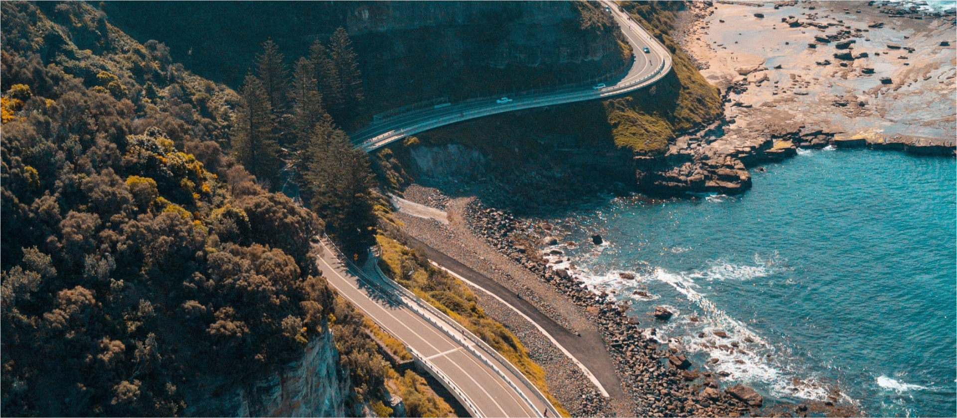An aerial shot of a coastal highway curving around rocky cliffs overlooking a bay of blue water.