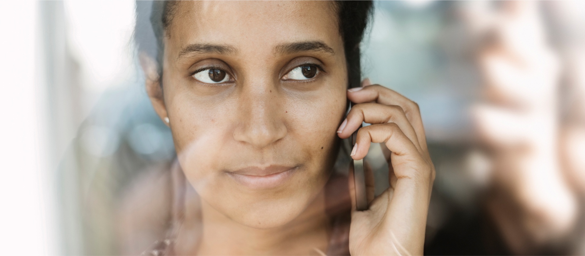 A woman speaking on a mobile device, as she looks out a window, the outside softly reflected in the pane of glass.