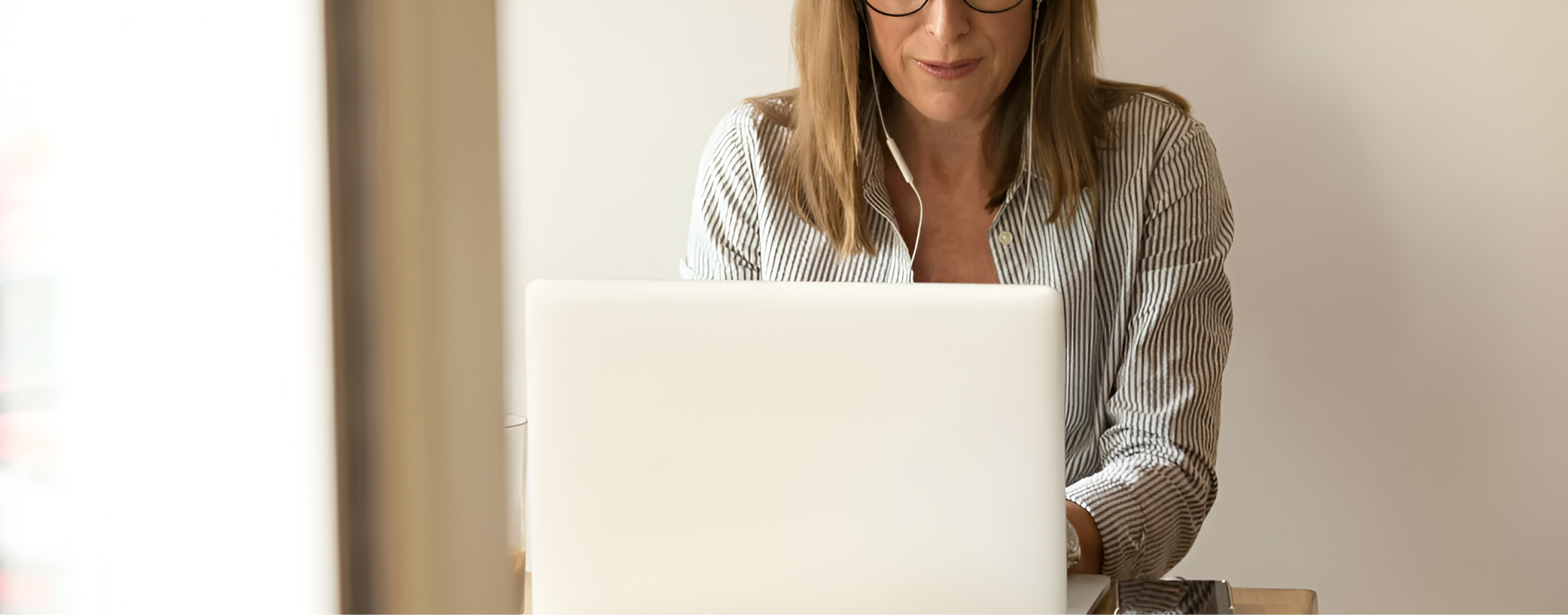 A woman in reading glasses with earphones plugged in, highly focused and typing away at a white laptop.