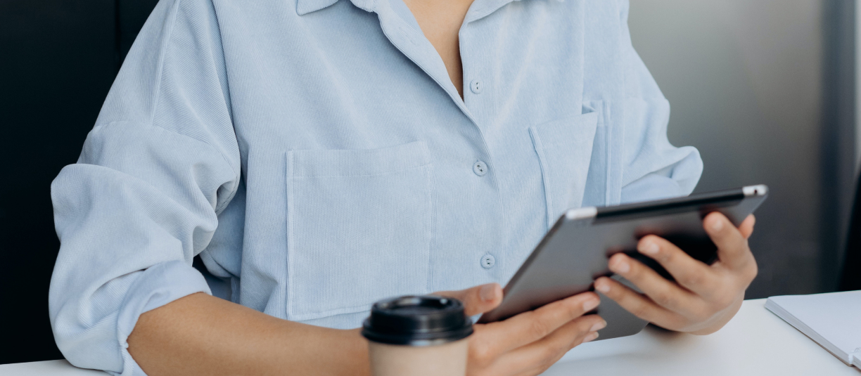 A professional woman in a light blue shirt reviews information on a digital tablet at her desk, with a coffee cup nearby.