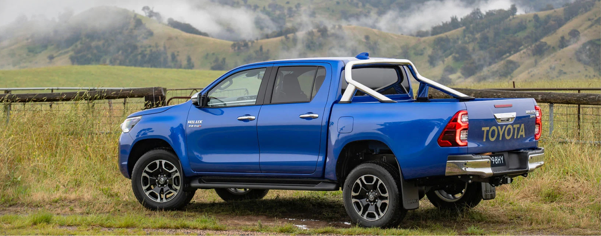 A blue Toyota HiLux parked facing a grassy paddock amongst misty mountains, in the Australian countryside.