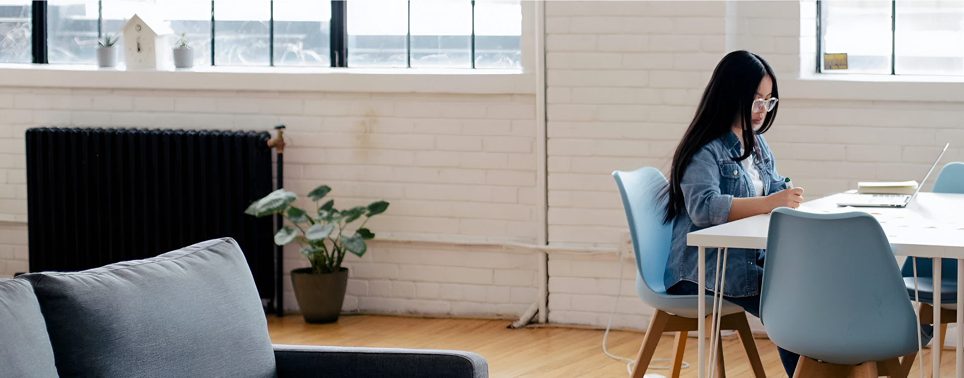 A woman sits at a modern white dining room table in a brightly-lit studio apartment, focusing on some paperwork with a laptop open in front of her.
