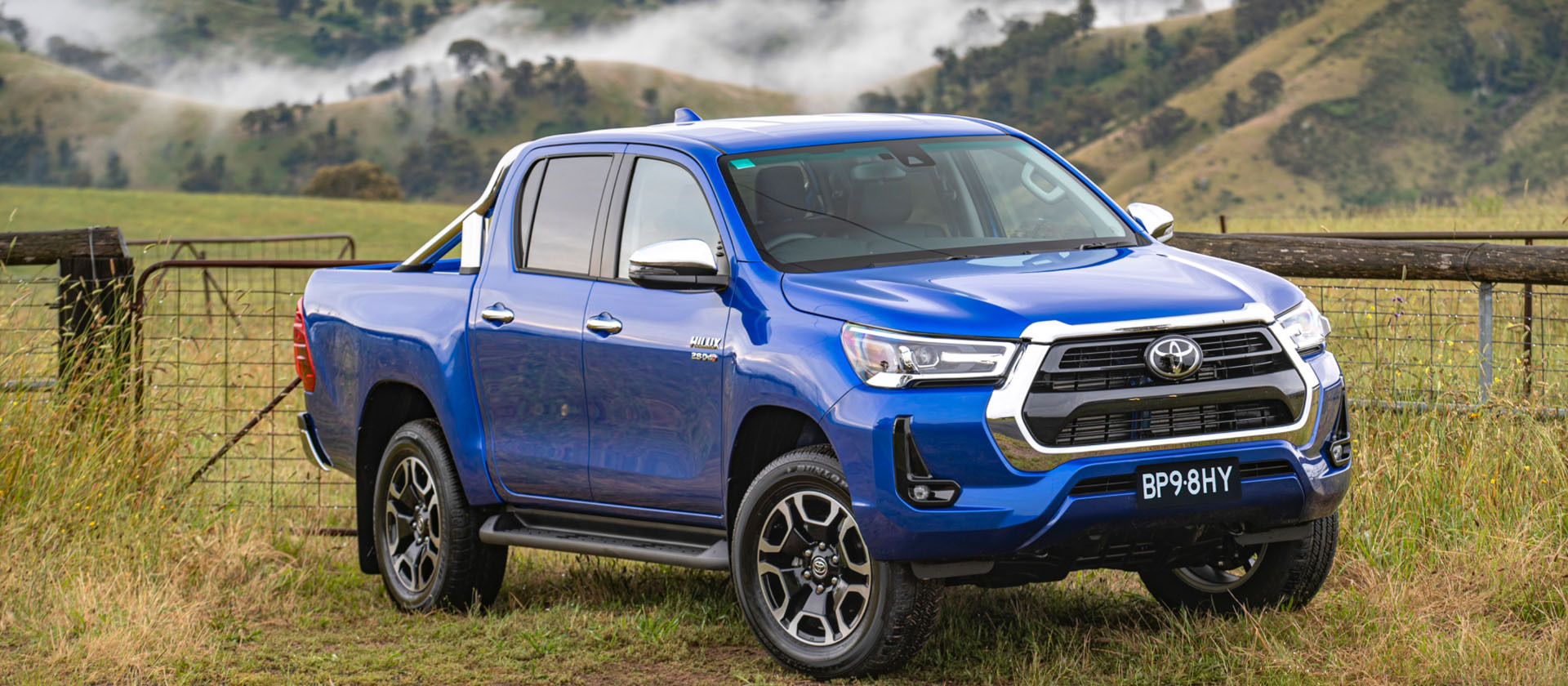 A blue Toyota HiLux parked in front of a grassy paddock amongst misty mountains, in regional Australia.