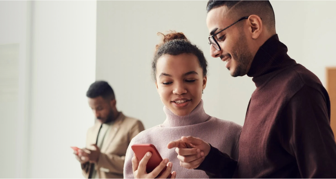 A man and a woman standing together side by side, both looking at the screen of a red smartphone being held up by the woman.