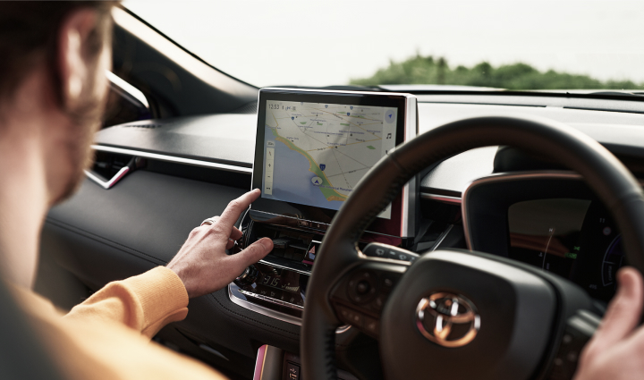A man drives a Toyota Corolla Cross, the view of the interior showing him reaching over to the vehicle’s built-in navigation on the multimedia display.