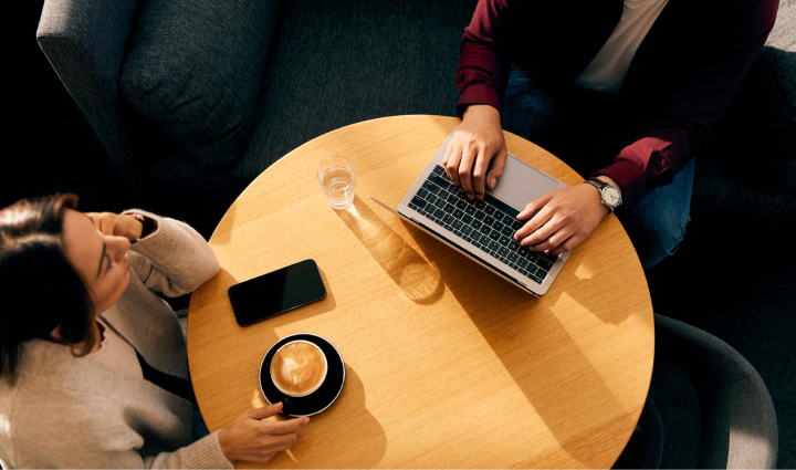 A birds eye view of two women at a circular table. The first woman has a mug of coffee in her hand with her other elbow resting on the table. The other woman typing away on her laptop with a glass of water beside her.