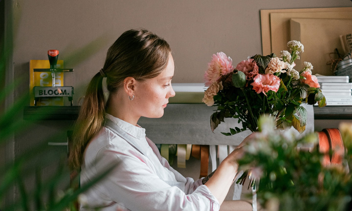 A woman doing floristry on a bunch of freshly-picked pink and cream-coloured flowers in a flower shop.