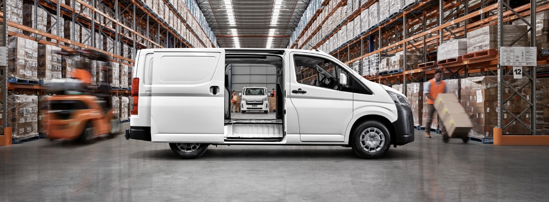 A white Toyota HiAce parked sideways in a storage warehouse. Its centre doors stand open, giving a view right through the side of the vehicle further into the warehouse, where another Toyota HiAce sits facing forward in the distance.