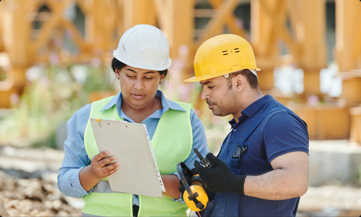 Man and woman wearing hardhats at a construction site, closely inspecting a clipboard.