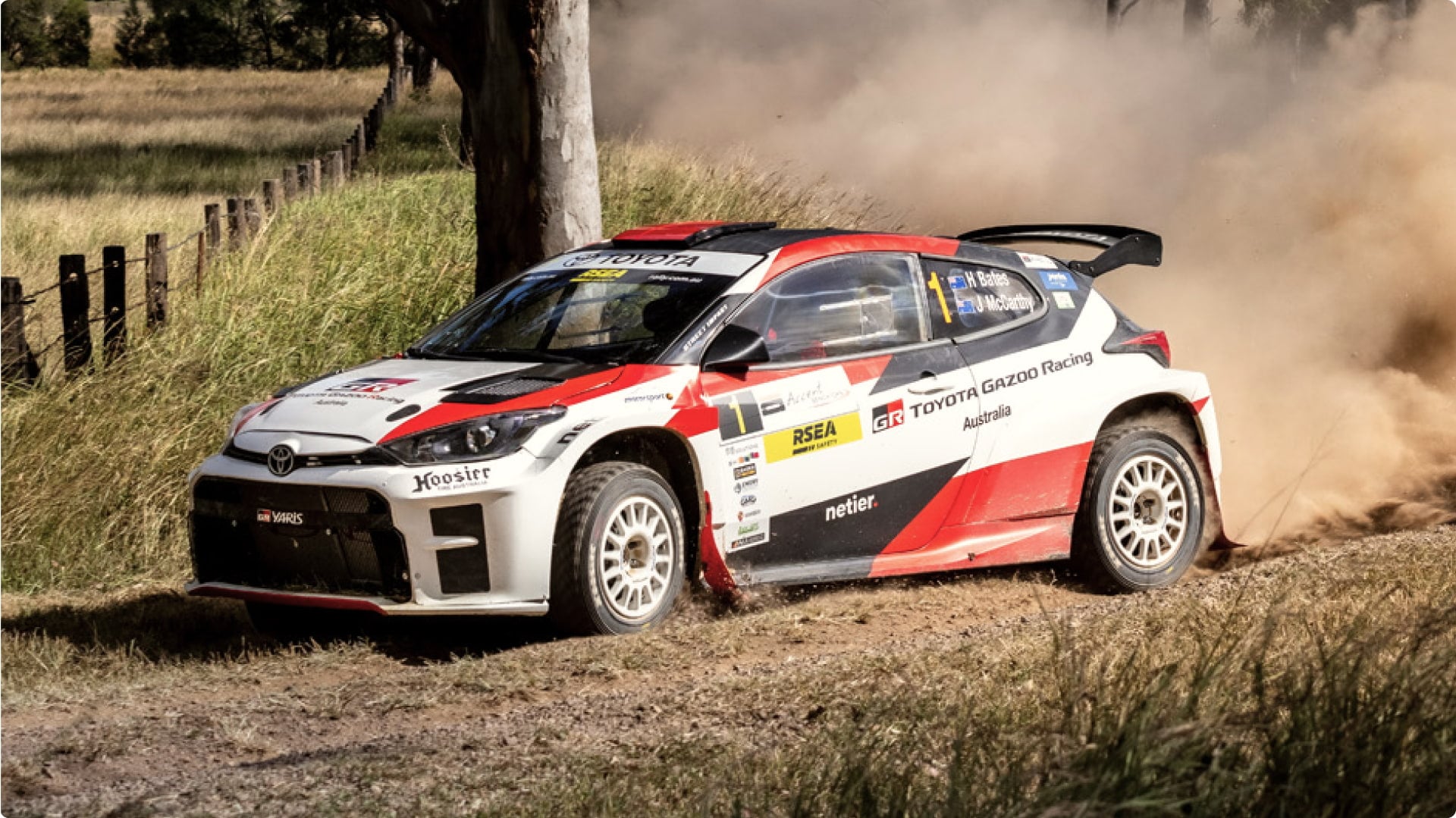 A white 2021 Toyota A R C G R Yaris A P 4 rally car at the Netier National Capital Rally, in red and black racing livery with sponsor logos. It's cornering a track bend surrounded by paddocks.