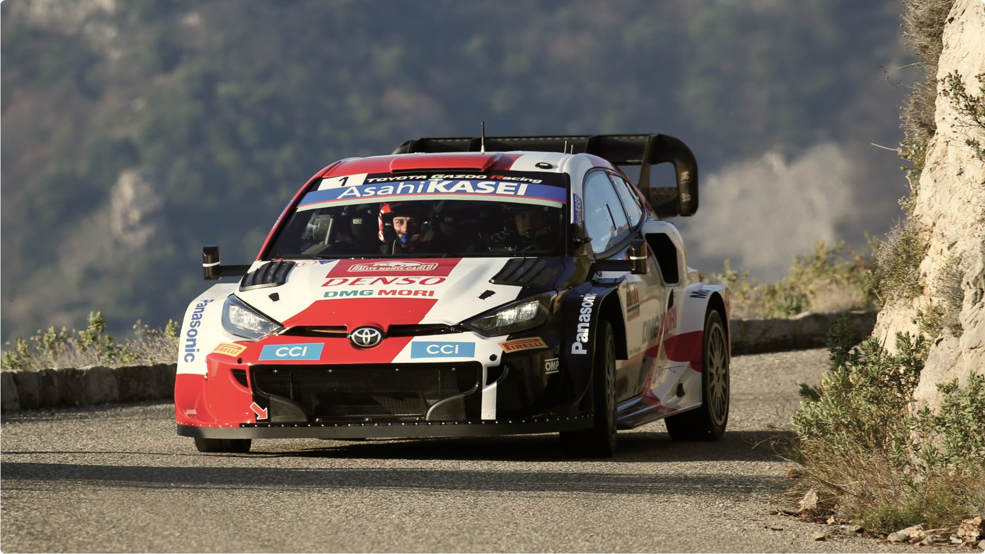 A 2022 Toyota W R C G R Yaris Rally1 in racing livery, driving along a road overlooking a cliff, with a tree-lined rockface visible in the background.