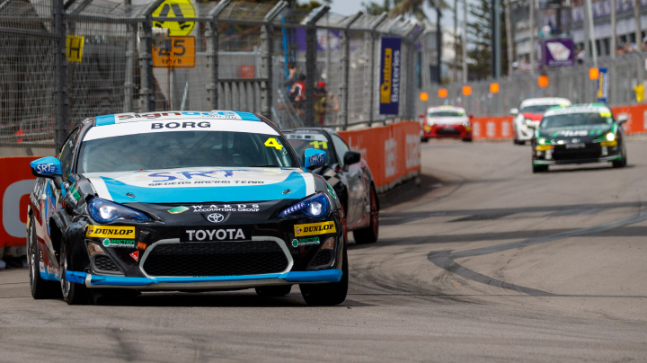 A Toyota G R 86 in blue and black racing livery leads other competitors at the Newcastle 500. The track is lined with tall steel fencing.
