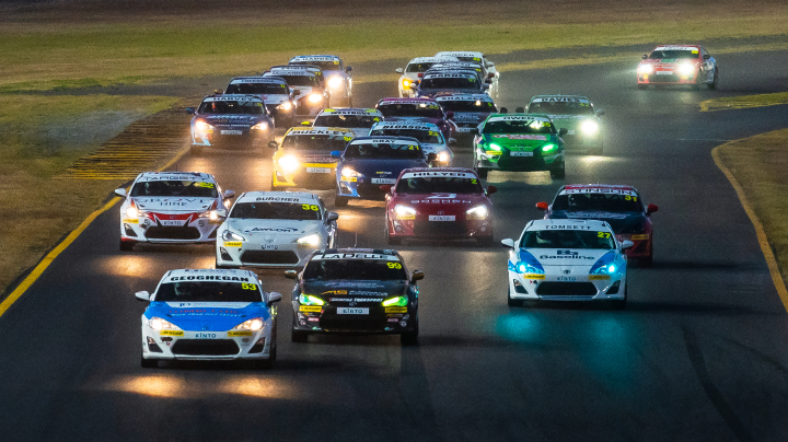 A large group of Toyota G R 86s in racing livery, competing at the Sydney Supernight, their headlights reflected in the track surface.