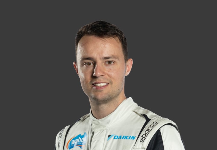 A headshot of professional T G R A driver Joshua Anderson in a white, black and light blue racing suit against a grey background.