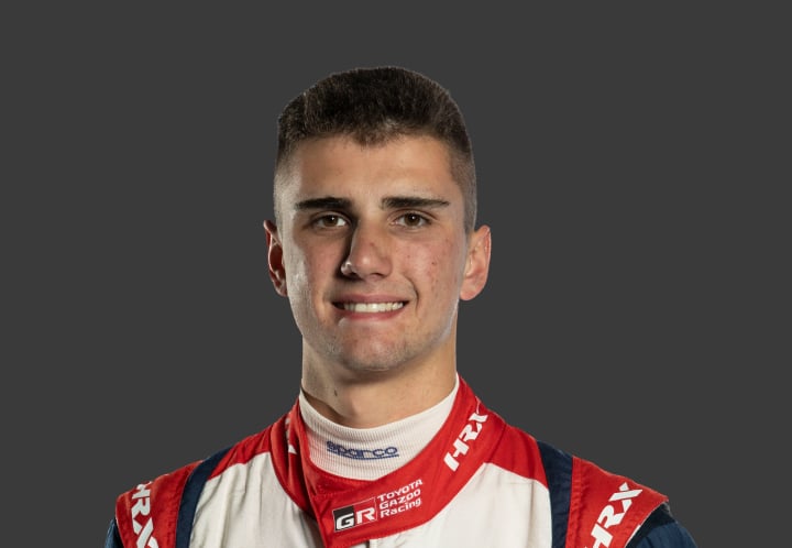A headshot of professional T G R A driver Cooper Barnes in a red, white and black racing suit against a grey background.