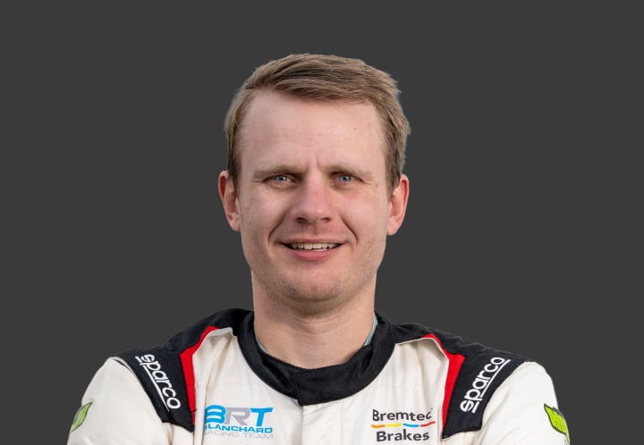 A headshot of professional T G R A driver Harrison Blanchard in a white, black and red racing suit against a grey background.