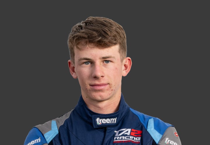 A headshot of professional T G R A driver Tyler Cheney in a racing suit of grey and blues against a grey background.