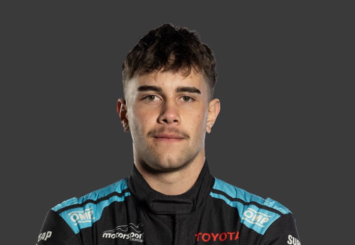 A headshot of professional T G R A driver Kade Davey in a black and light blue racing suit against a grey background.