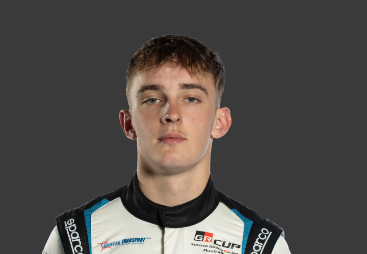 A headshot of professional T G R A driver Toby Dvorak in a white, black and light bkue racing suit against a grey background.