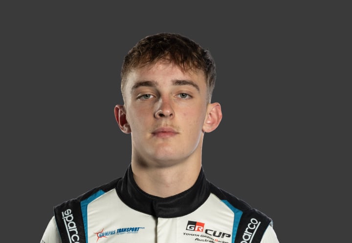A headshot of professional T G R A driver Toby Dvorak in a white, black and light bkue racing suit against a grey background.