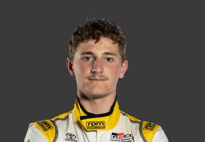 A headshot of professional T G R A driver Lachlan Evennett in white and yellow racing suit against a grey background.