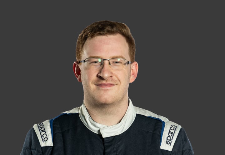 A headshot of professional T G R A driver Lachlan Gibbons, in a dark navy and white racing suit against a grey background.