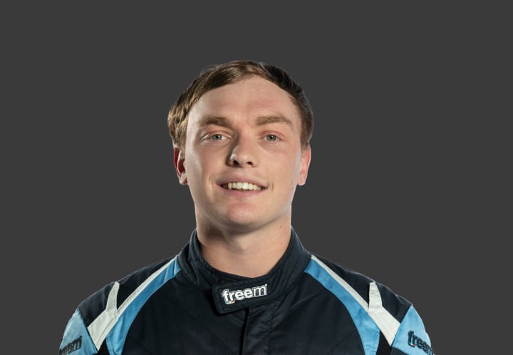 A headshot of professional T G R A driver Hayden Hume in a dark navy blue, light blue and white racing suit against a grey background.