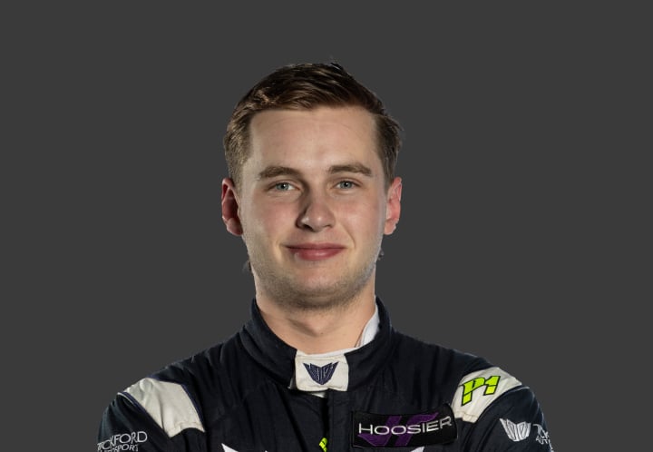 A headshot of professional T G R A driver Charlie Nash in a black, green and white racing suit against a grey background.