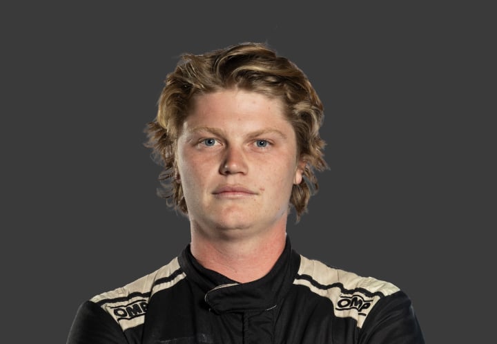 A headshot of professional T G R A driver Charlie Parker in a black and white racing suit against a grey background.