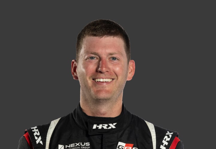 A headshot of professional T G R A driver Shane Roberts in a black, red and white racing suit against a grey background.
