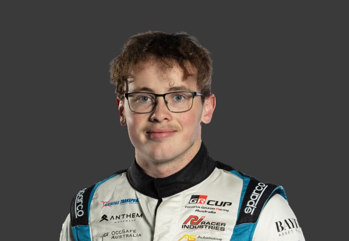 A headshot of professional T G R A driver Ben Stewart in a blue and white racing suit against a grey background.